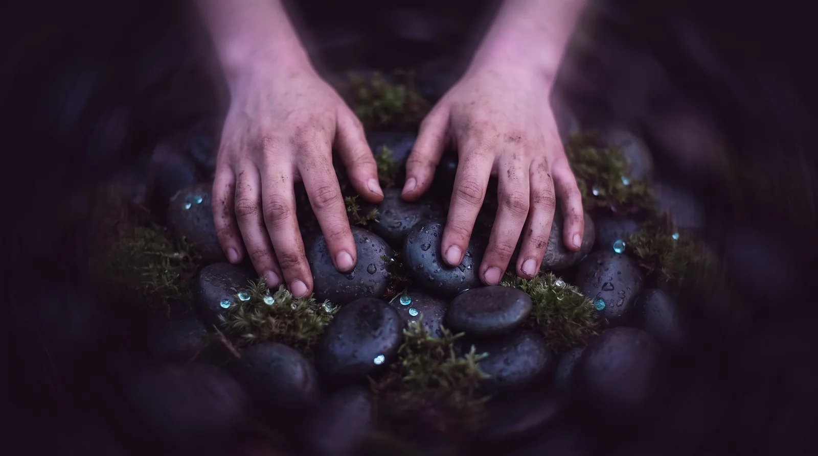 Two hands pressing gently into dark river stones and moss, fingers traced with lavender and rose light against near-black shadow.