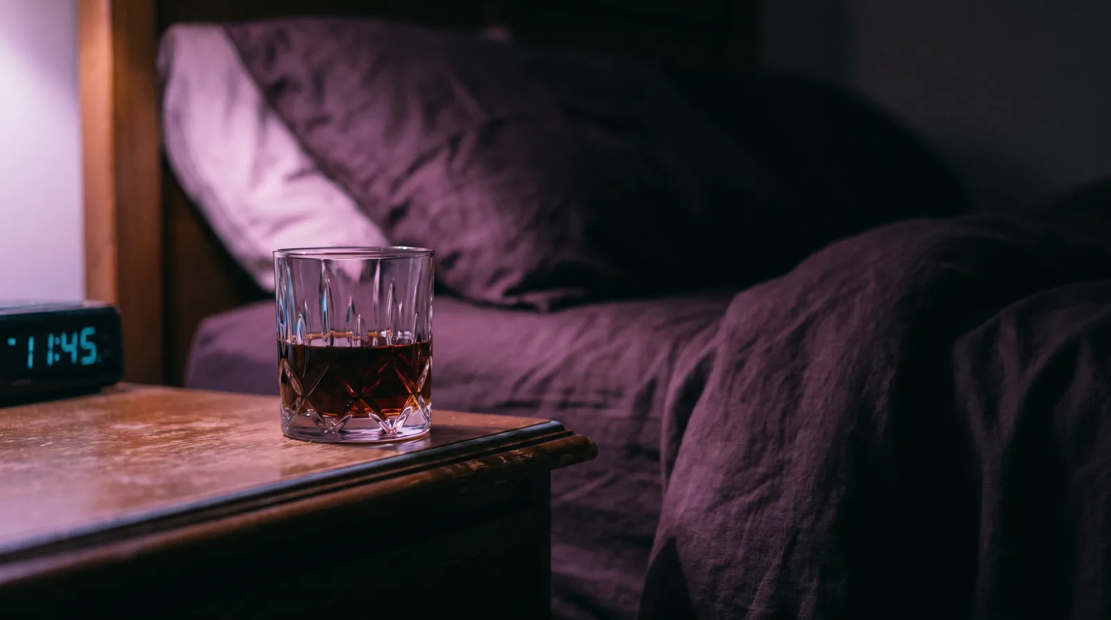 A glass of amber liquid on a dark nightstand, rimmed in rose and lavender light, bedding fading into near-black.