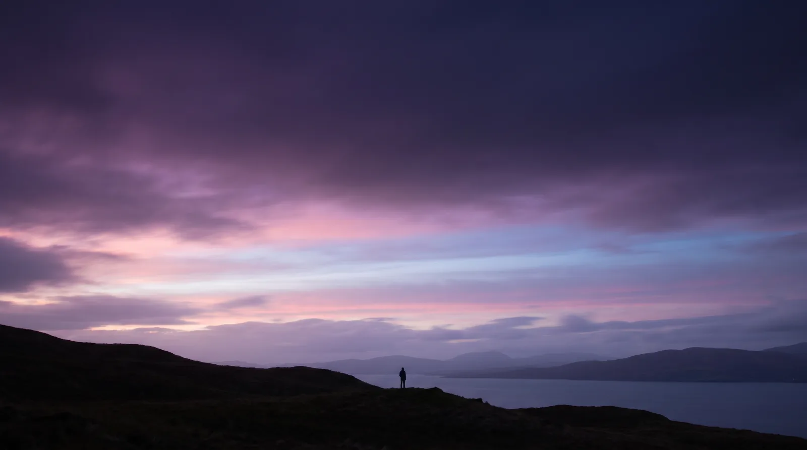 A tiny silhouetted figure on a hillside beneath a vast twilight sky banded with lavender, rose and aqua afterglow.