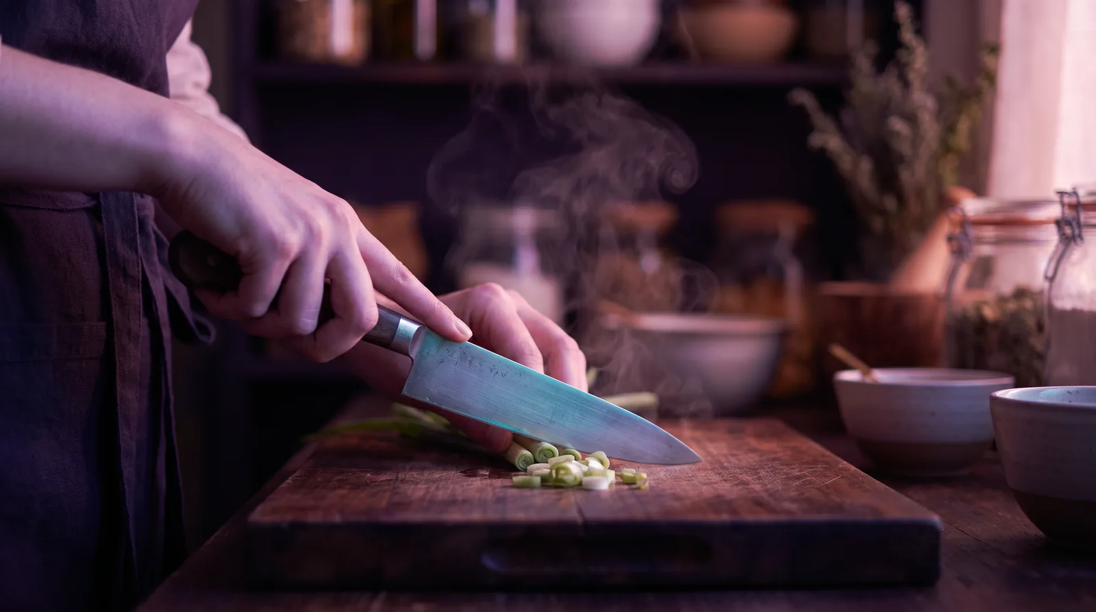Hands slicing vegetables on a wooden board in soft lavender and rose light, steam rising past a dark aubergine background with aqua glints on the knife.