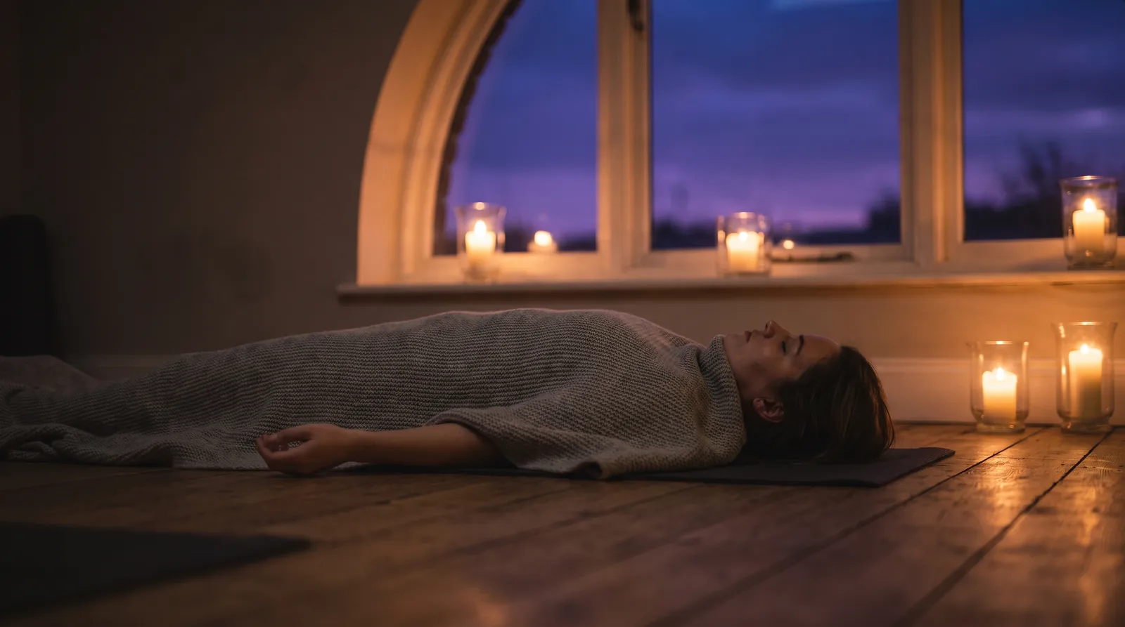 A person resting in savasana under a blanket in a dim candle-lit studio.