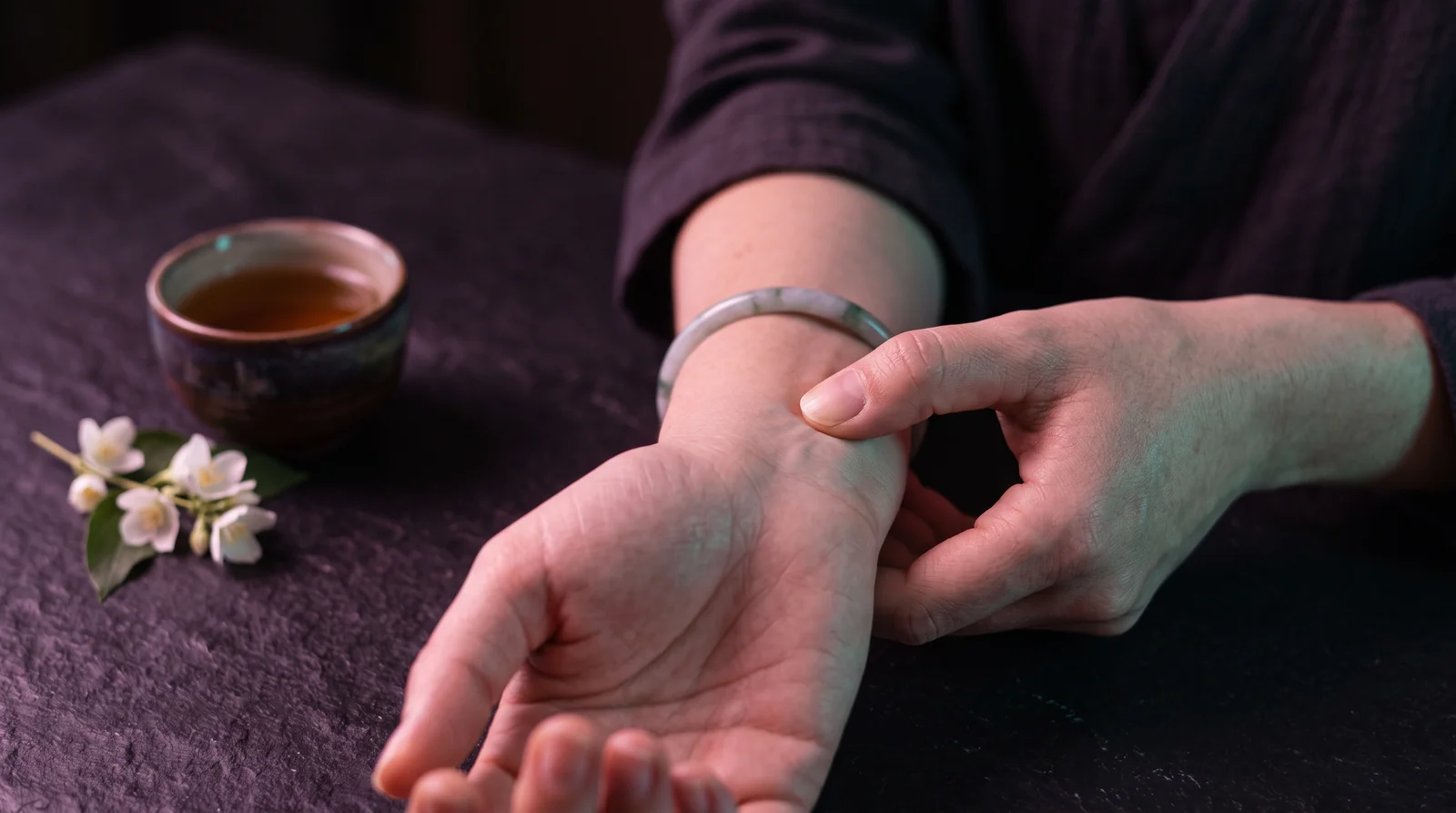 A thumb pressing gently into the inner wrist of the other hand in soft violet light.