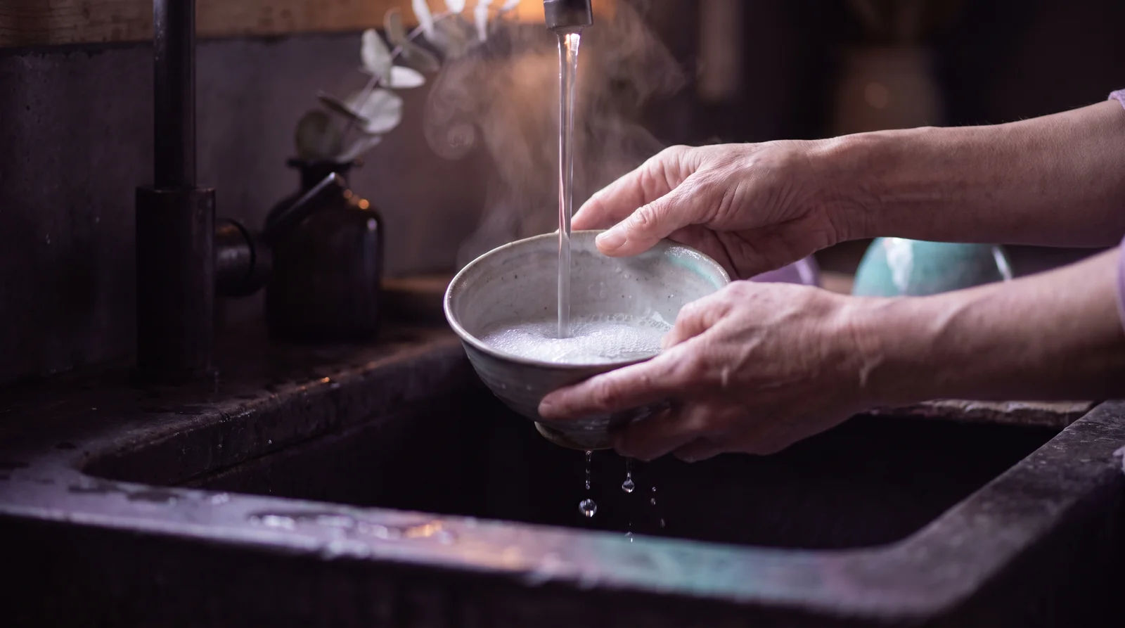 Hands rinsing a pale ceramic bowl under a stream of water in a dim kitchen, soap film catching lavender, rose and aqua light against deep aubergine shadow.