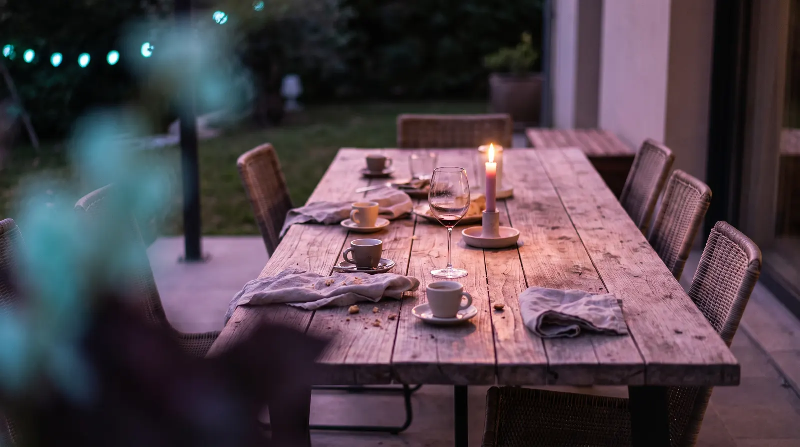 A long wooden table after a meal, empty cups and a low candle glowing rose and lavender against deep aubergine dusk.