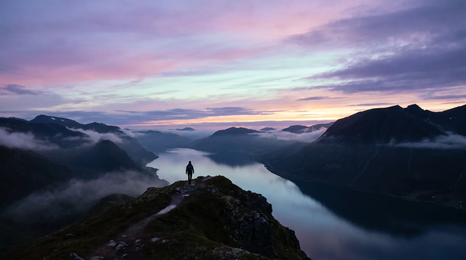 A lone figure on a quiet mountain path above a mirrored fjord at dusk, the sky washed in lavender, rose and aqua over deep aubergine peaks.