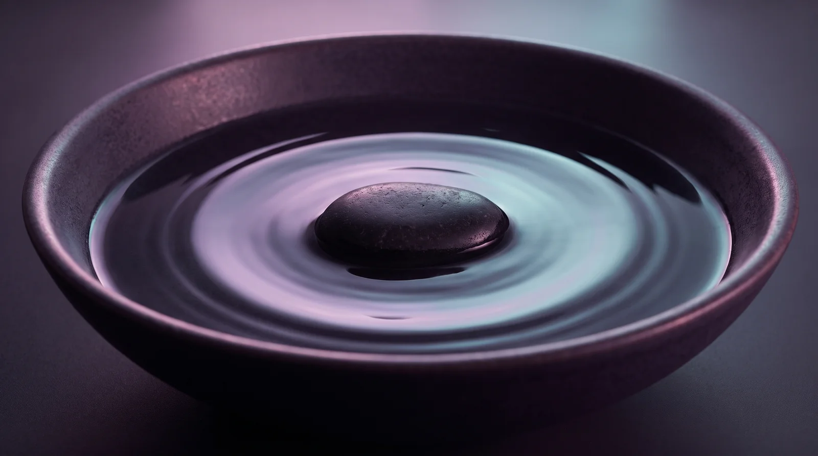 A single smooth dark stone at the centre of still water in a shallow ceramic bowl, one ripple spreading outward in lavender and aqua light.