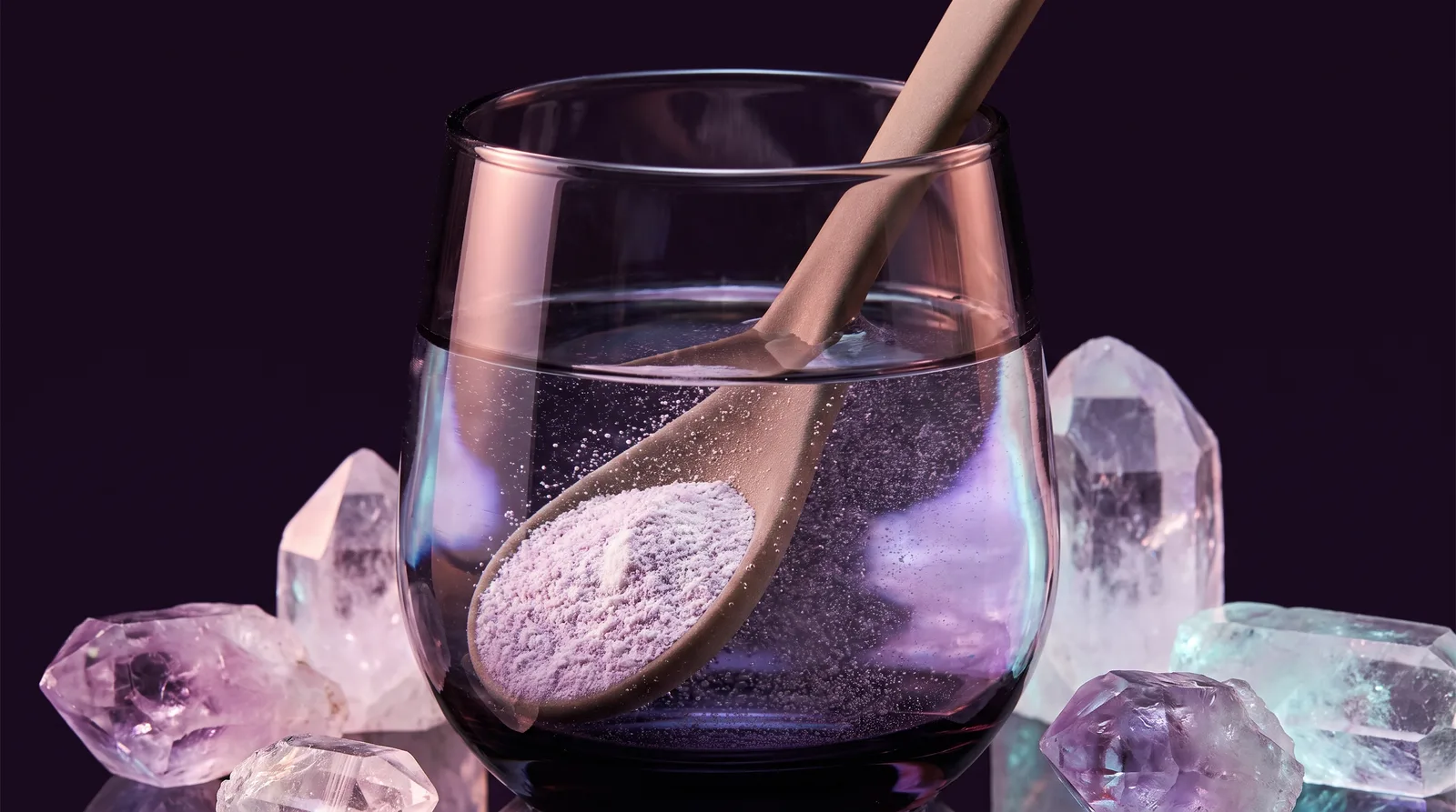 Pale mineral crystals and powder dissolving into a dark glass of water lit with lavender and aqua glow.