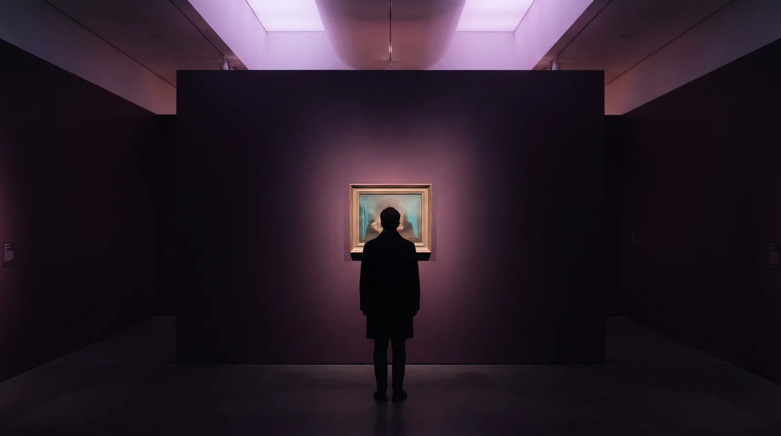 A lone figure in a dark gallery contemplating a single softly glowing painting on a near-black wall.
