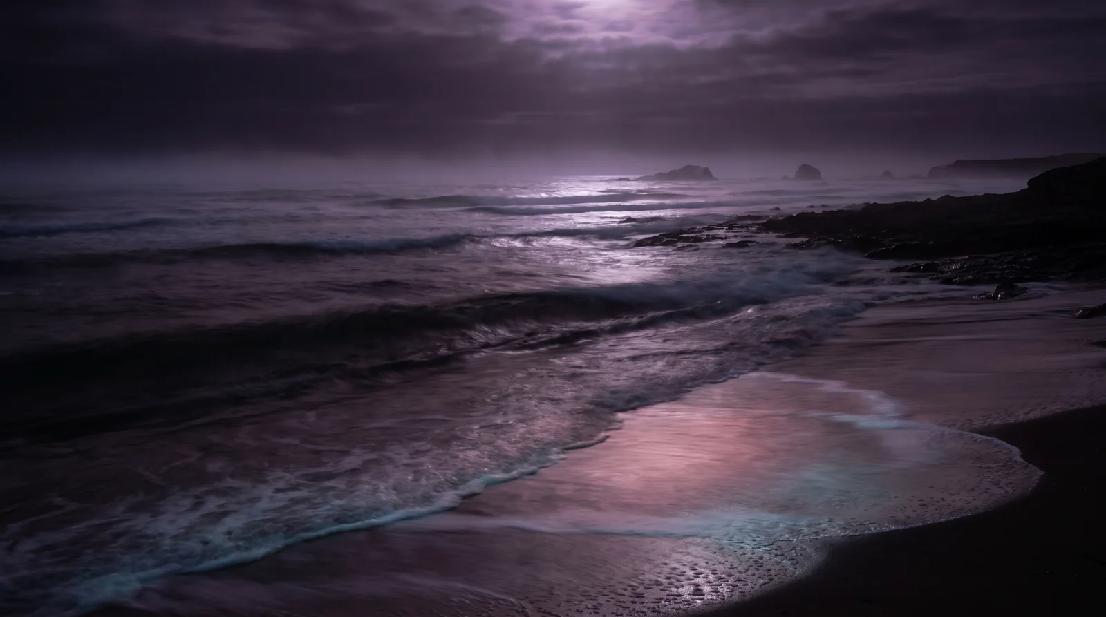 Slow silvery waves rolling toward a dark shore under a lavender night sky.