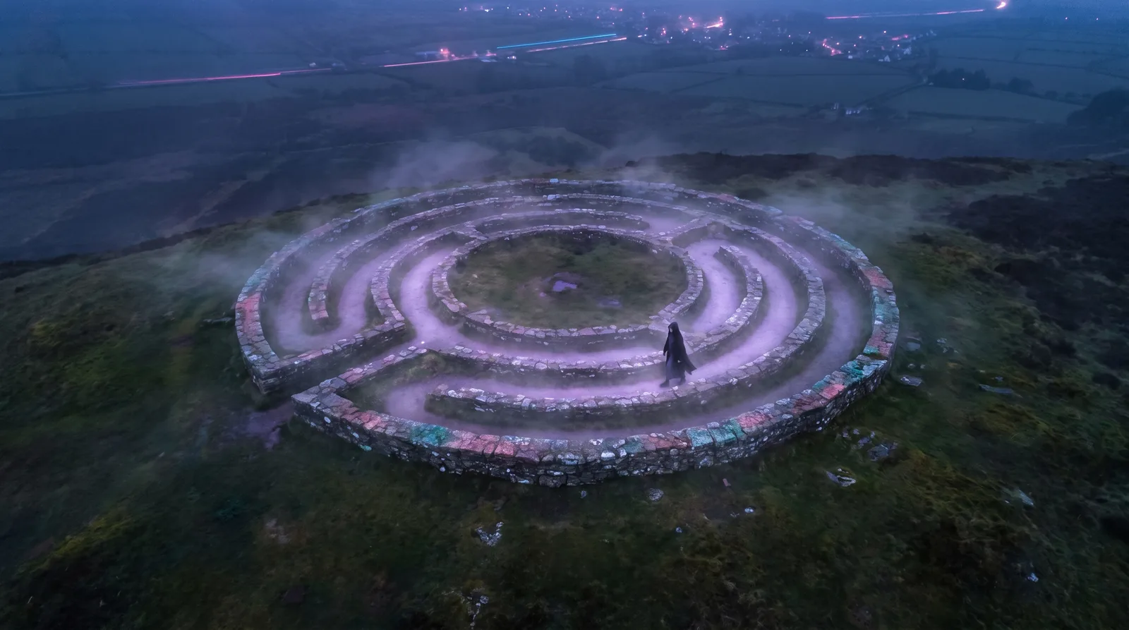An ancient stone labyrinth seen from above at dusk, its single winding path glowing lavender through drifting mist as a lone figure walks.