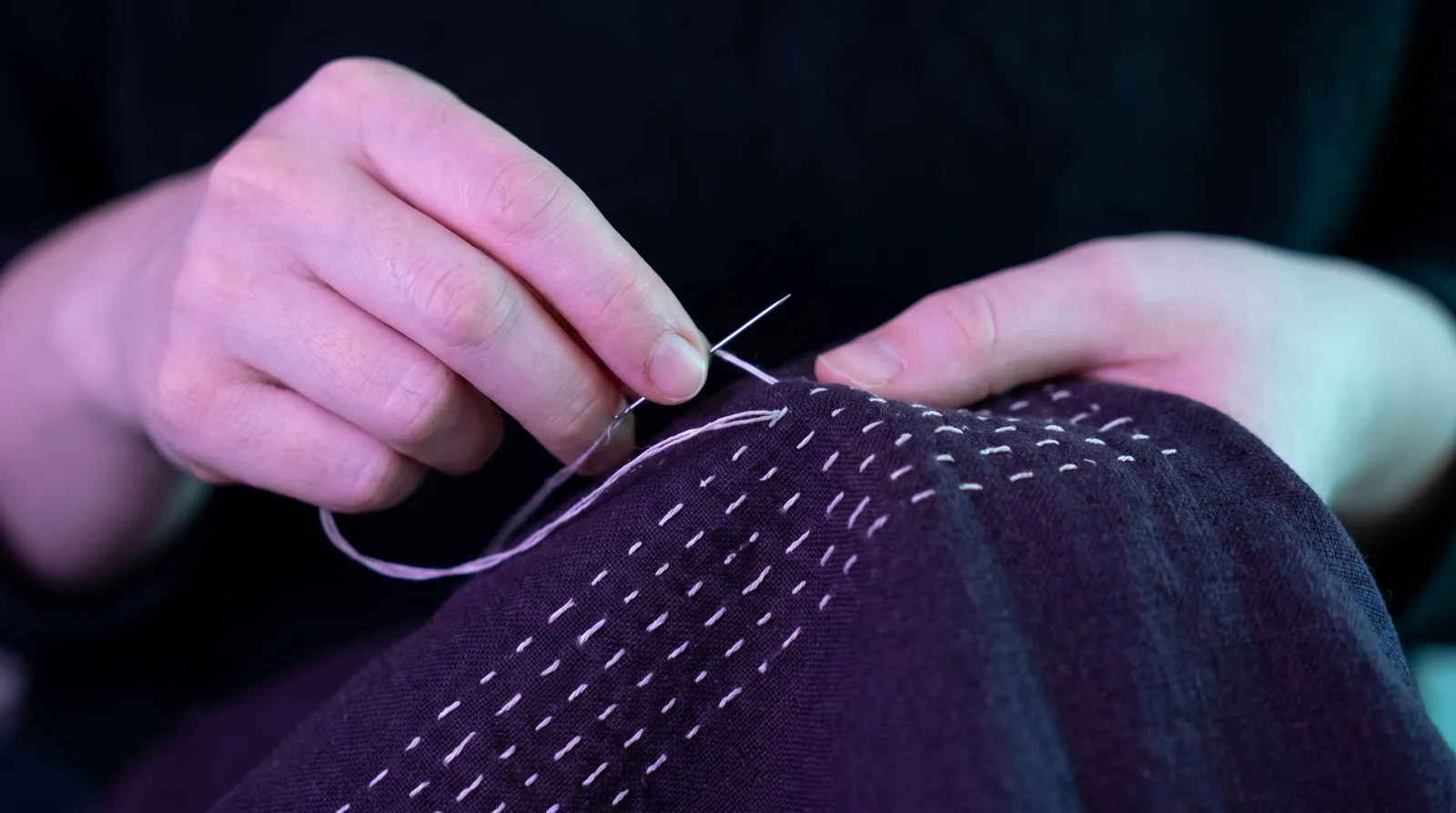 Hands guiding a needle through deep indigo cloth, a row of pale running stitches catching soft lavender light.