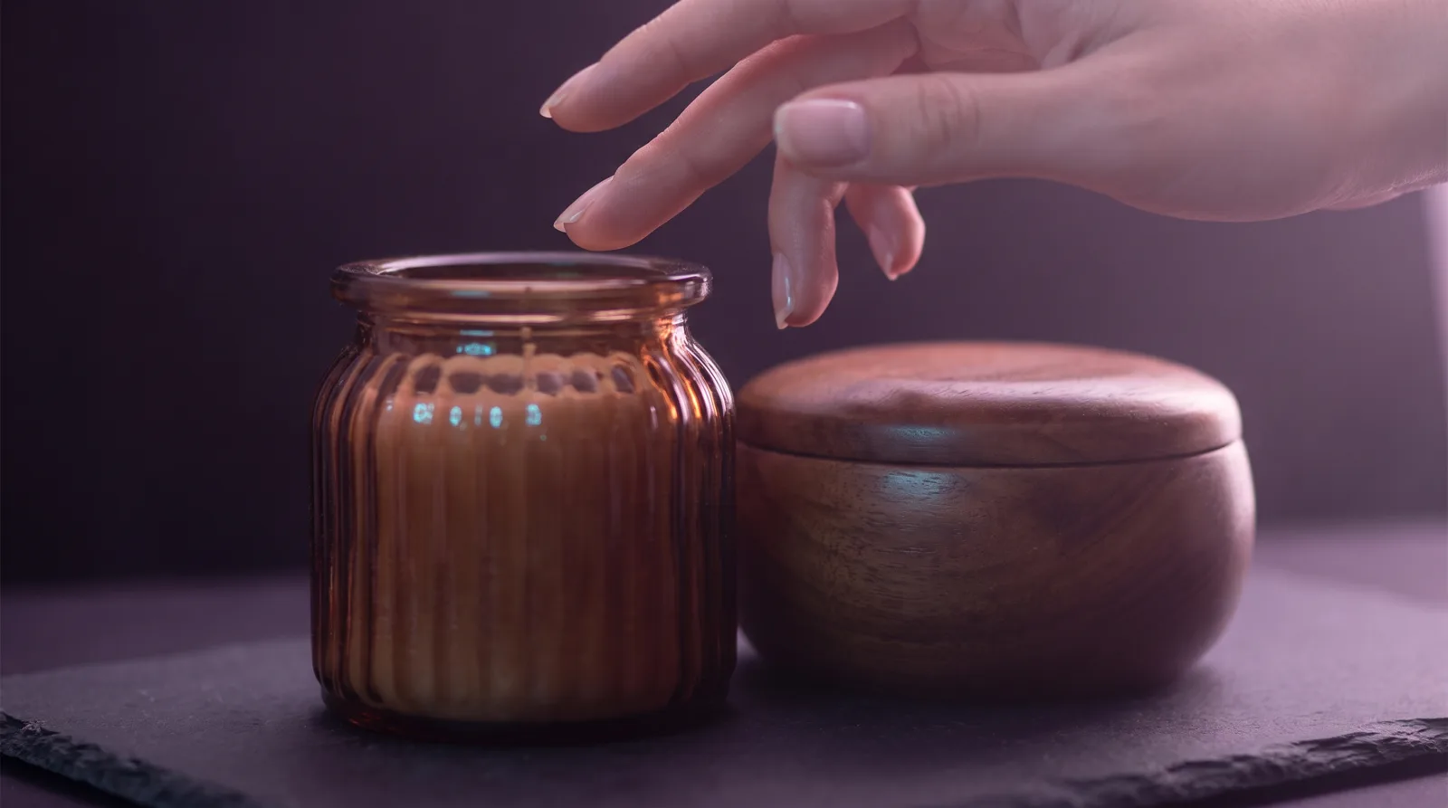Fingertips hovering above a ridged glass jar on a dark surface, rim-lit in lavender and rose against deep aubergine shadows.
