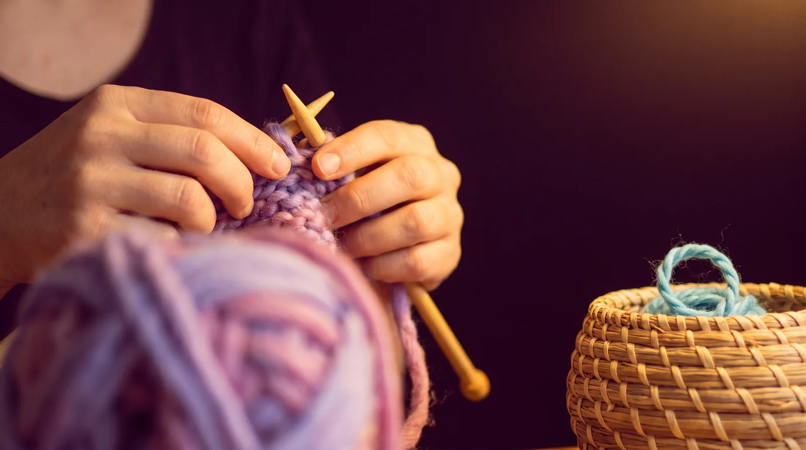 Hands knitting with wooden needles and soft lavender yarn in warm light against a deep aubergine background.