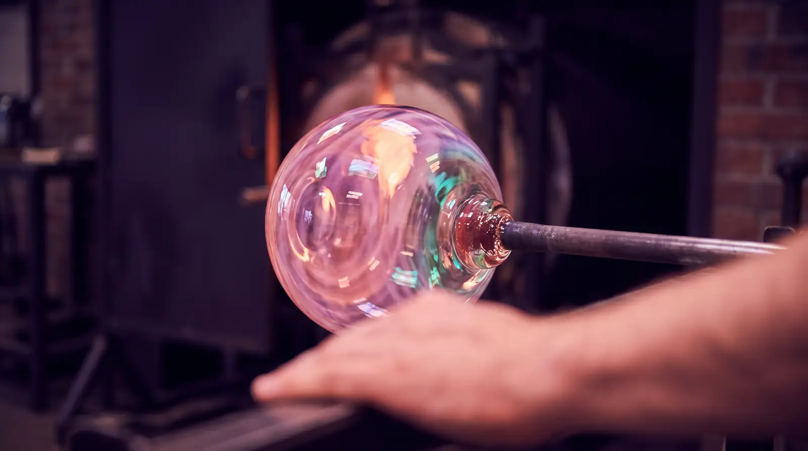 A molten glass orb glowing rose and lavender at the end of a blowpipe, turning slowly against deep aubergine workshop darkness.