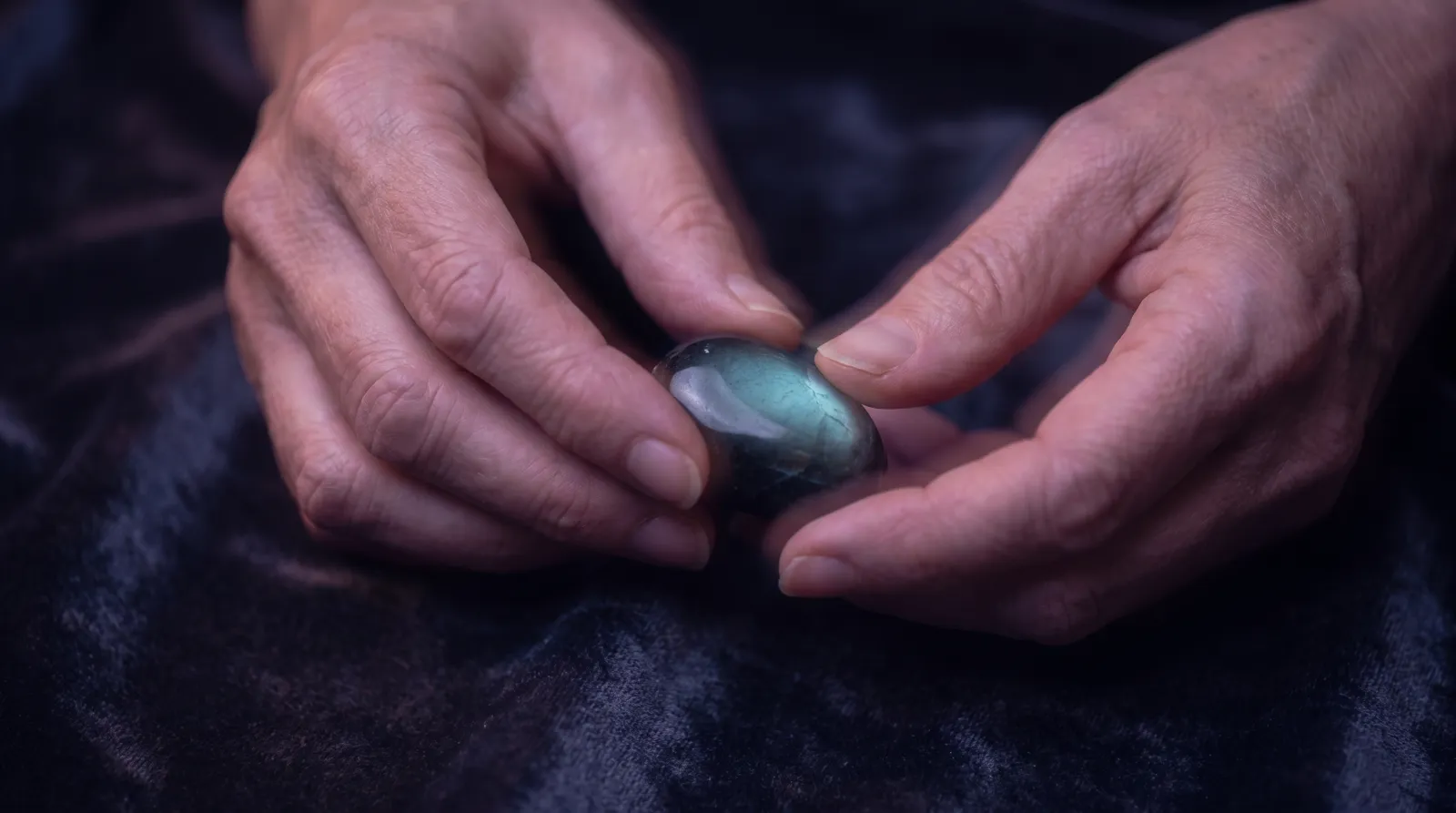 Hands rolling a smooth polished worry stone over dark velvet, rim-lit in lavender and rose with a soft aqua glow, in deep aubergine shadow.