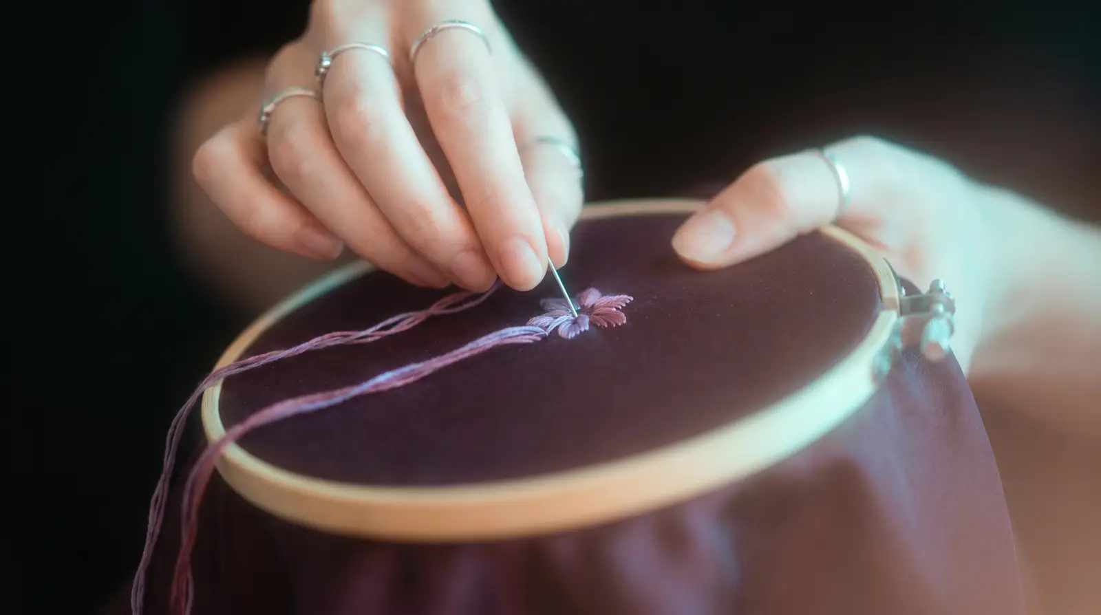 Hands embroidering silk in a round wooden hoop, a needle trailing lavender thread, lit by soft aqua light against deep aubergine darkness.
