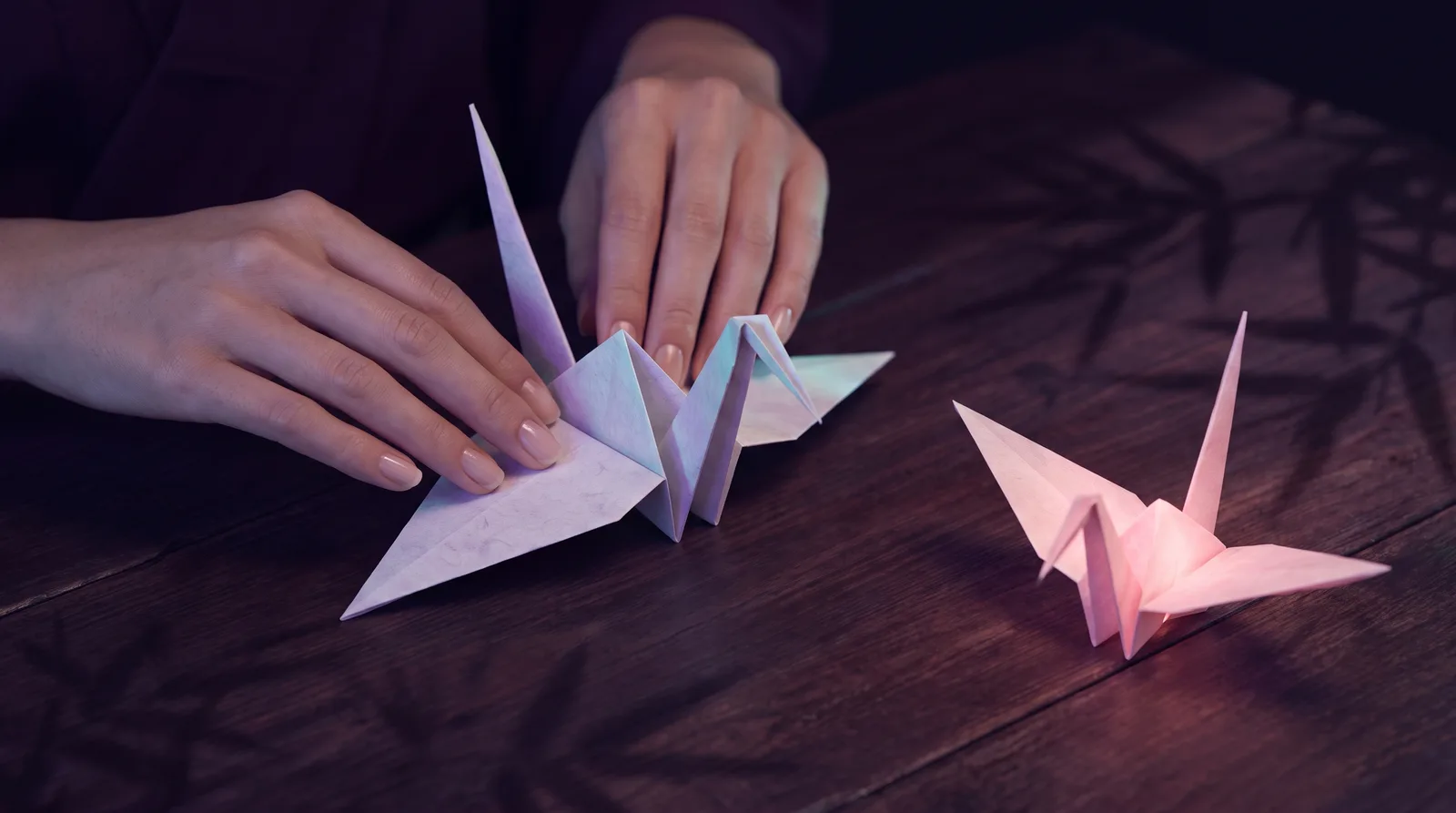 Hands creasing pale origami paper into a crane on a dark surface, edges catching soft aqua light.