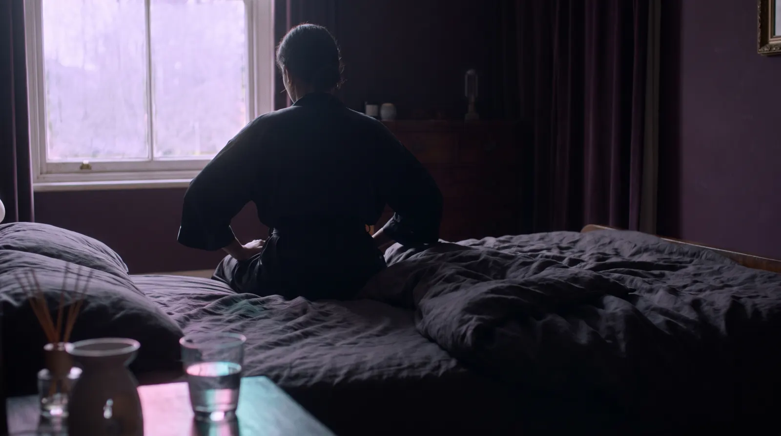 A person sitting on the edge of a bed at dawn, soft lavender light through the window.