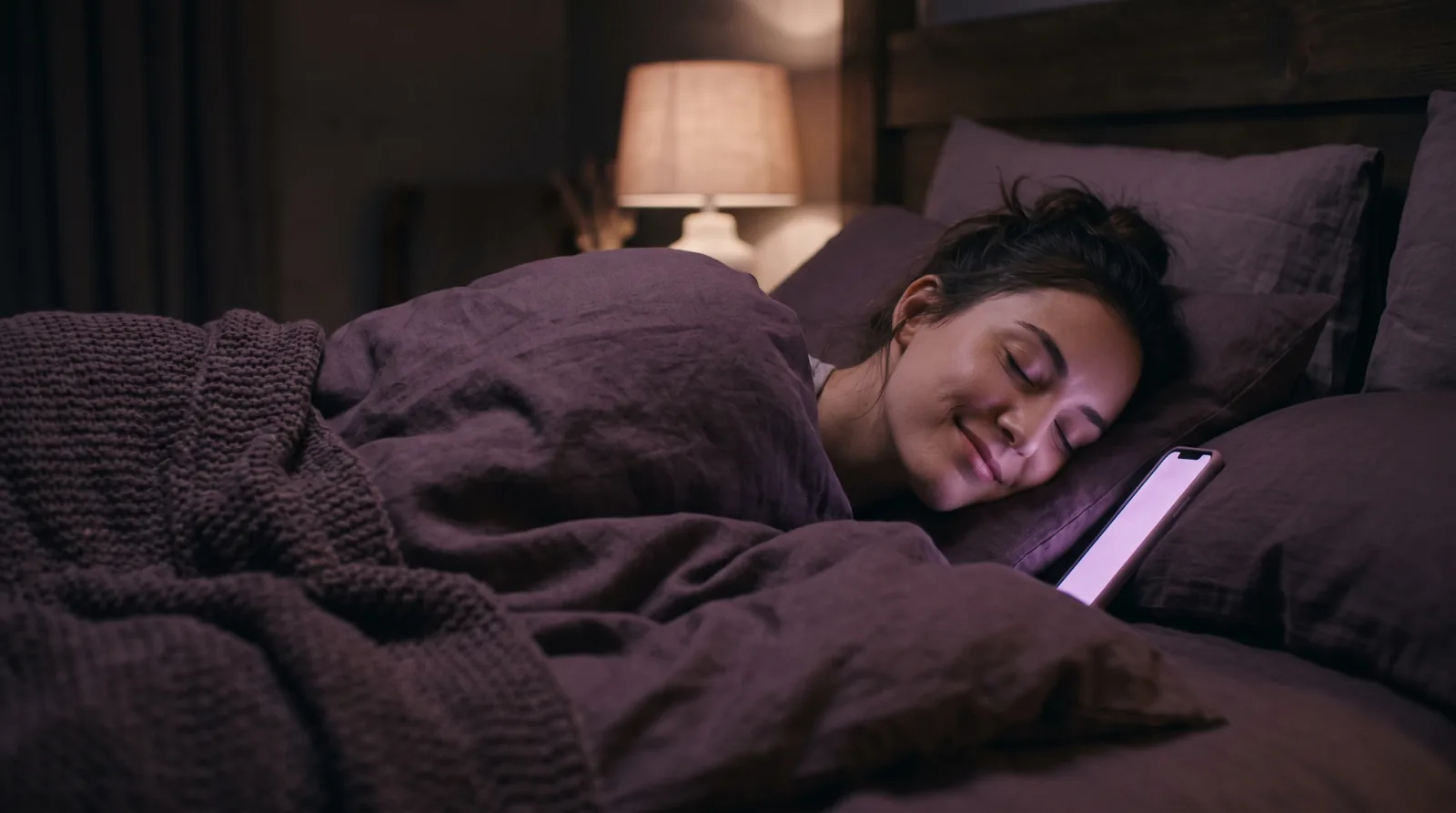 A woman drifting off in bed, her face softly lit by the calm lavender glow of her phone.