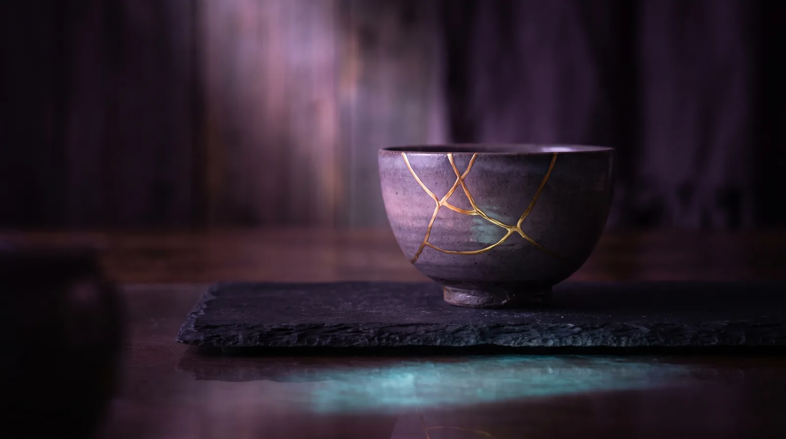 A dark ceramic bowl mended with luminous gold seams, resting on stone in soft lavender light.