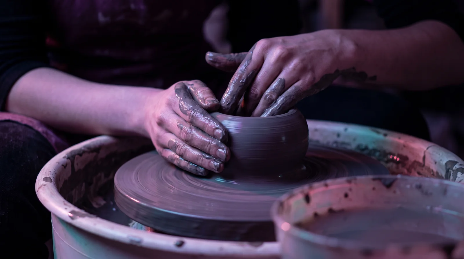 Hands shaping wet clay on a spinning wheel, lit in lavender and rose against deep aubergine shadow.