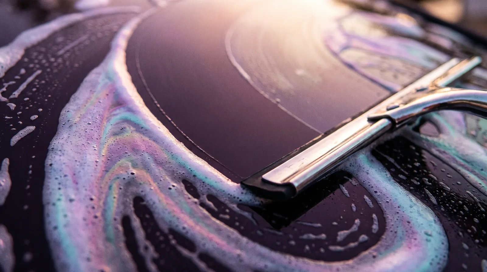 A squeegee pulling a clean arc through iridescent suds on dark glass.