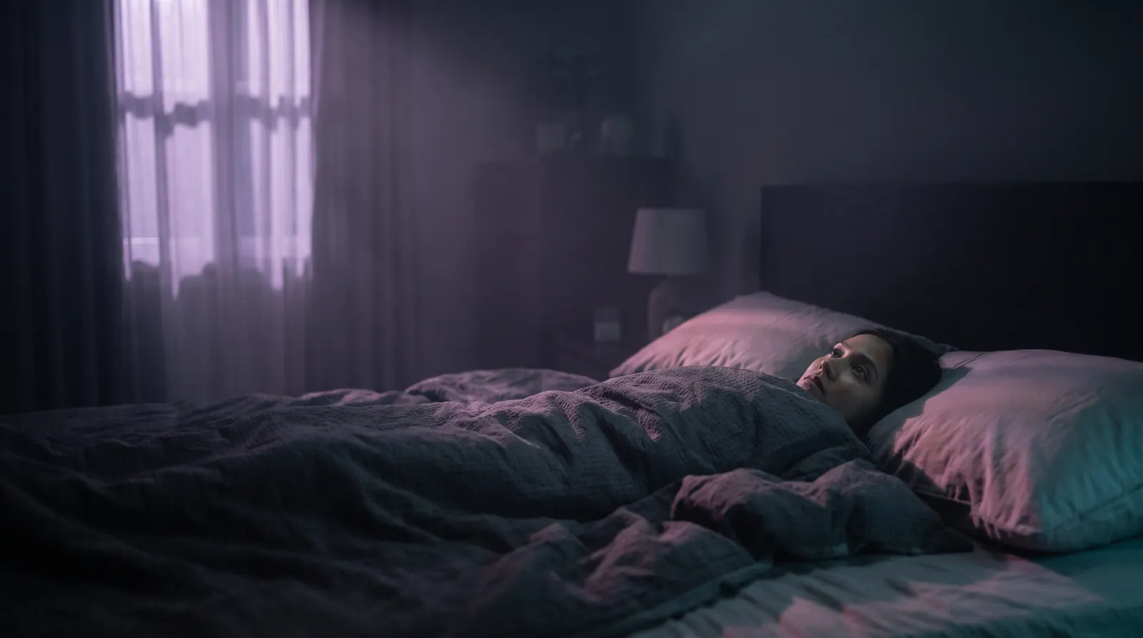 A person lying awake in bed at night in soft lavender moonlight.