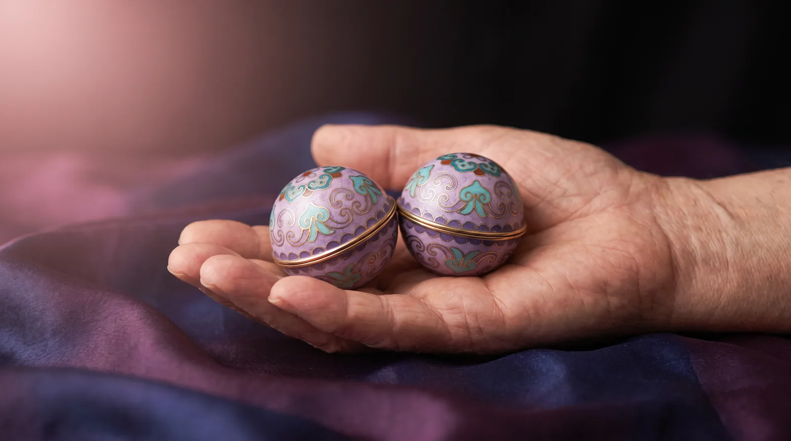 Two gleaming cloisonné baoding balls resting in an open palm, their lavender and aqua enamel catching soft rose light against deep aubergine shadow.