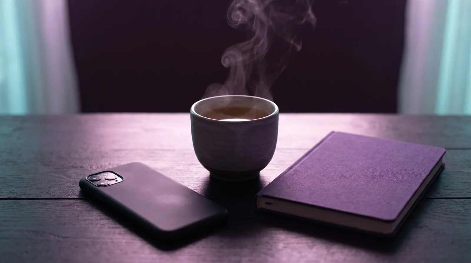 A smartphone lying face down on a dark table beside a steaming cup of tea and a closed notebook.