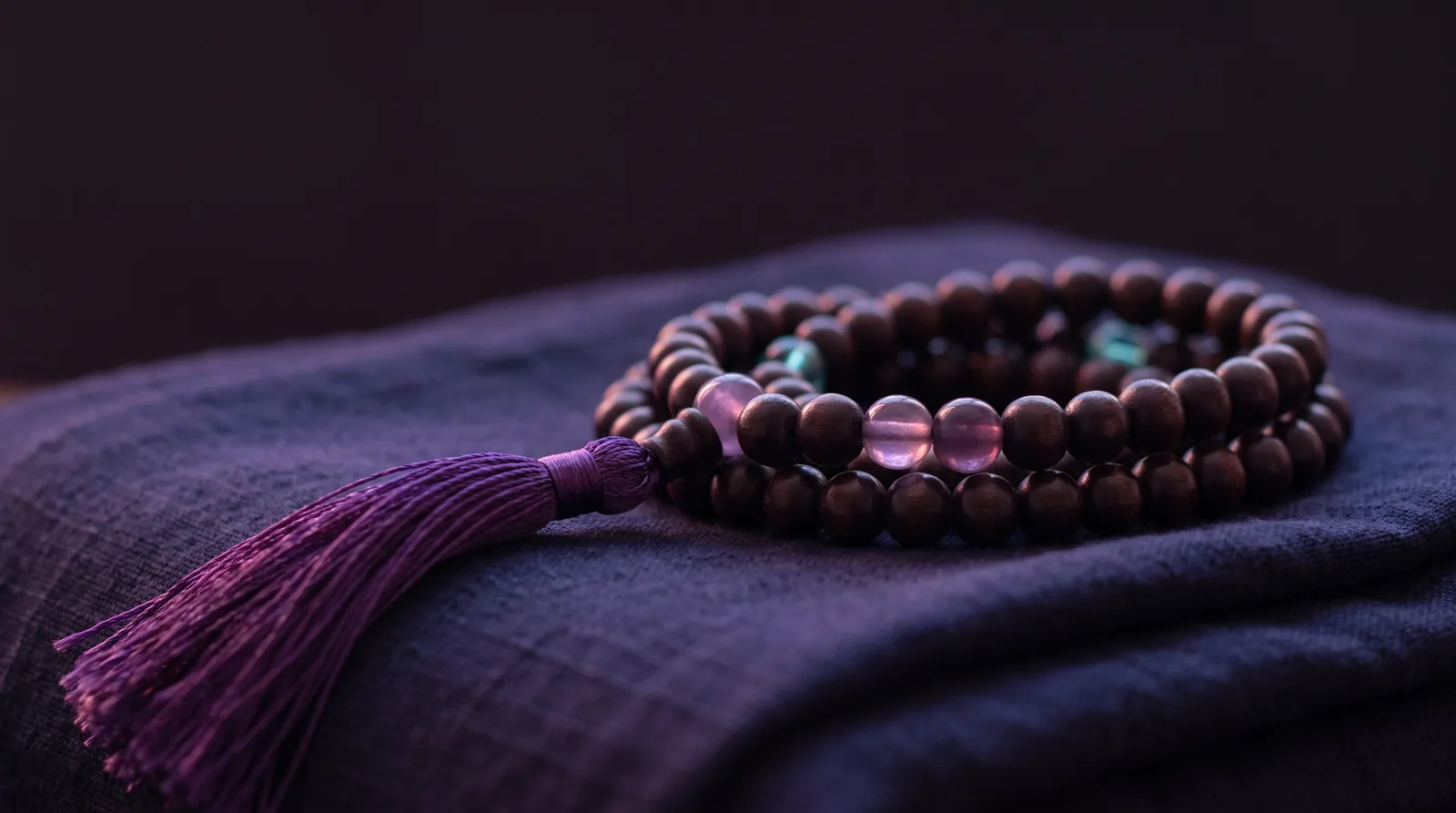 A strand of wooden mala beads with a silk tassel coiled on dark fabric, glowing softly in rose and lavender light.
