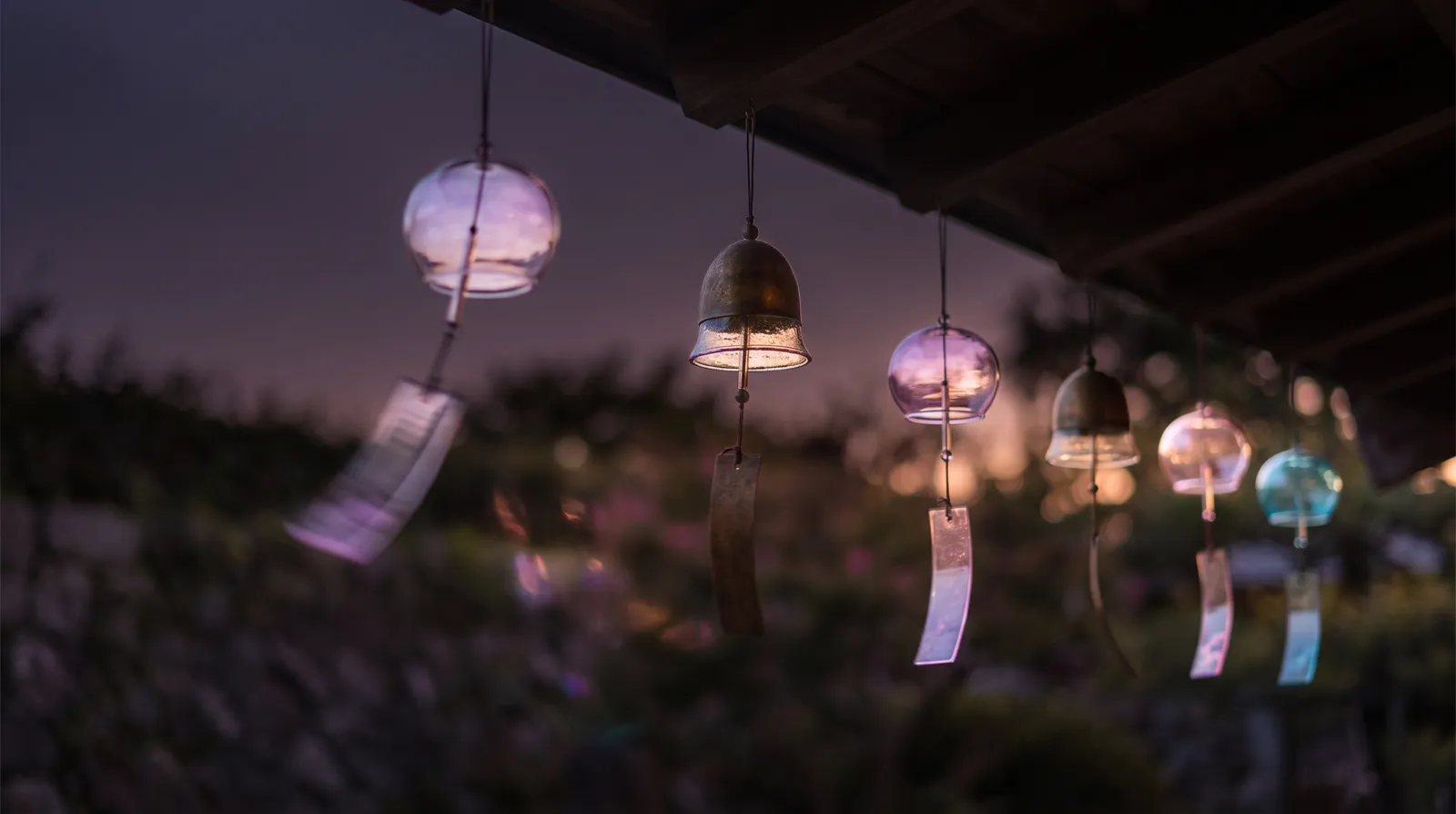 Glass and metal wind chimes hanging from a dark eave at dusk, one blurred mid-swing, lavender, rose and aqua light refracting against a deep aubergine gloom.