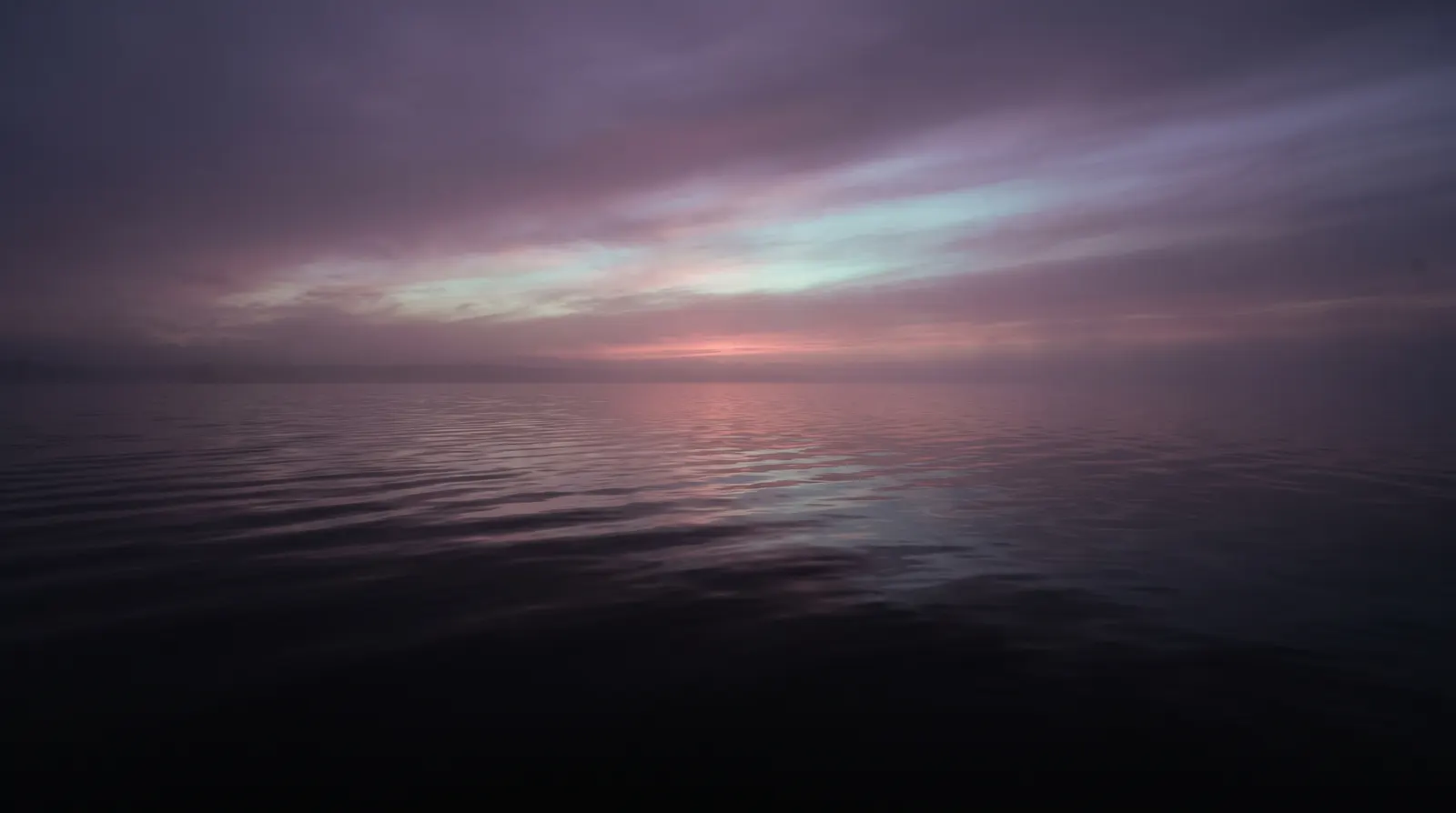 A vast calm water surface at dusk with slow ripples reflecting aqua and lavender light under a faint rose horizon.