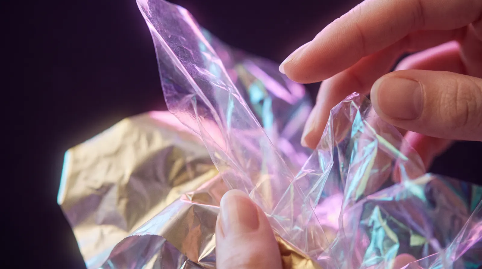 Fingertips holding crinkled translucent film, its crisp folds catching lavender, rose and aqua light against a deep aubergine background.