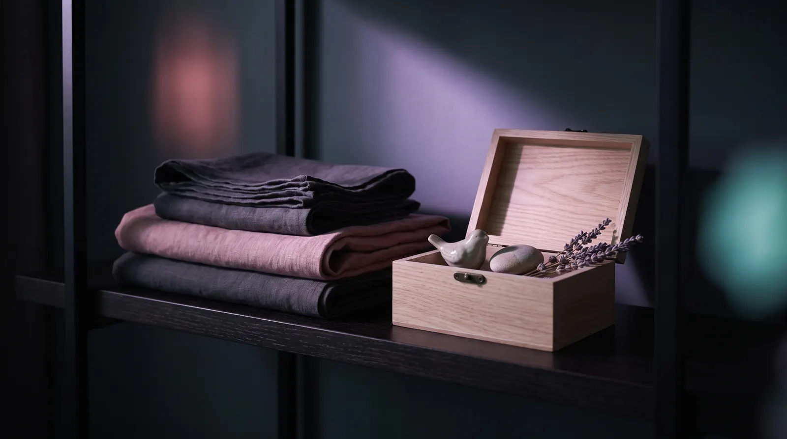 Neatly folded linens and a small wooden keepsake box on a dark minimalist shelf, lit by a soft beam of lavender light.
