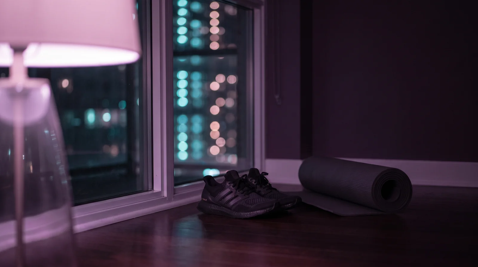 A pair of running shoes and a rolled yoga mat by a tall window at night, city lights blurred in aqua and rose against a deep aubergine room.
