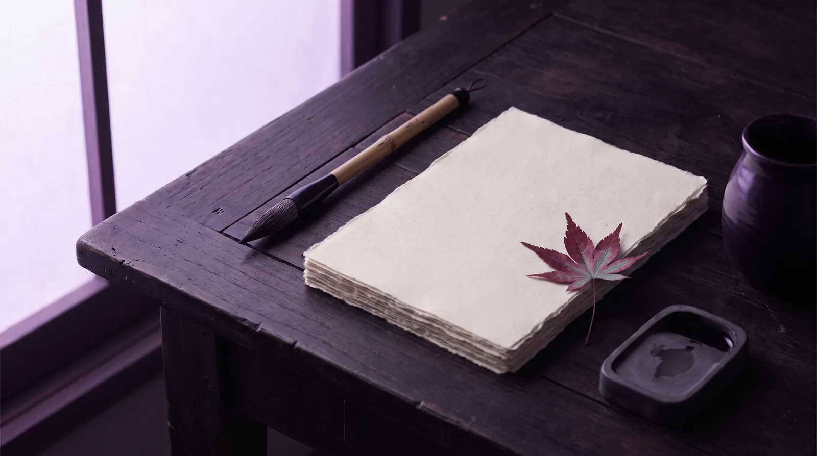 An ink brush resting beside blank paper and a single maple leaf on a desk lit by soft lavender light.