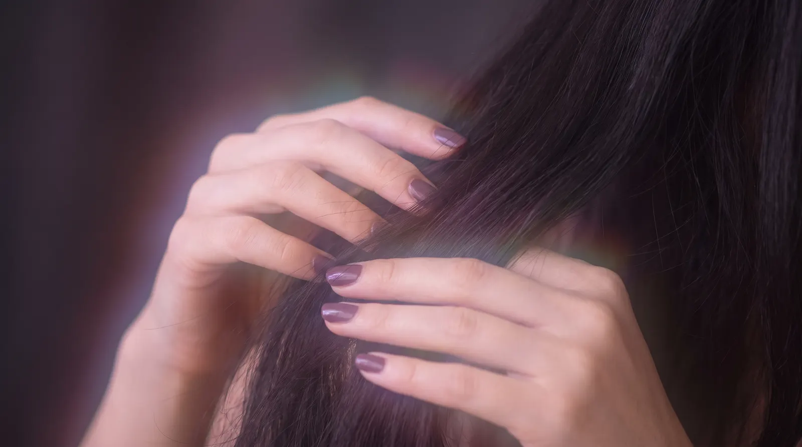 Hands gently running through long dark hair traced with lavender and rose light on a deep aubergine background.