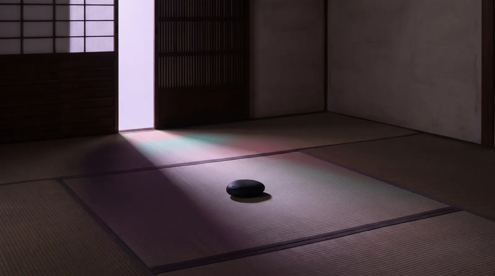 A nearly empty room with a single smooth stone on the floor and pale lavender light falling across deep shadow.